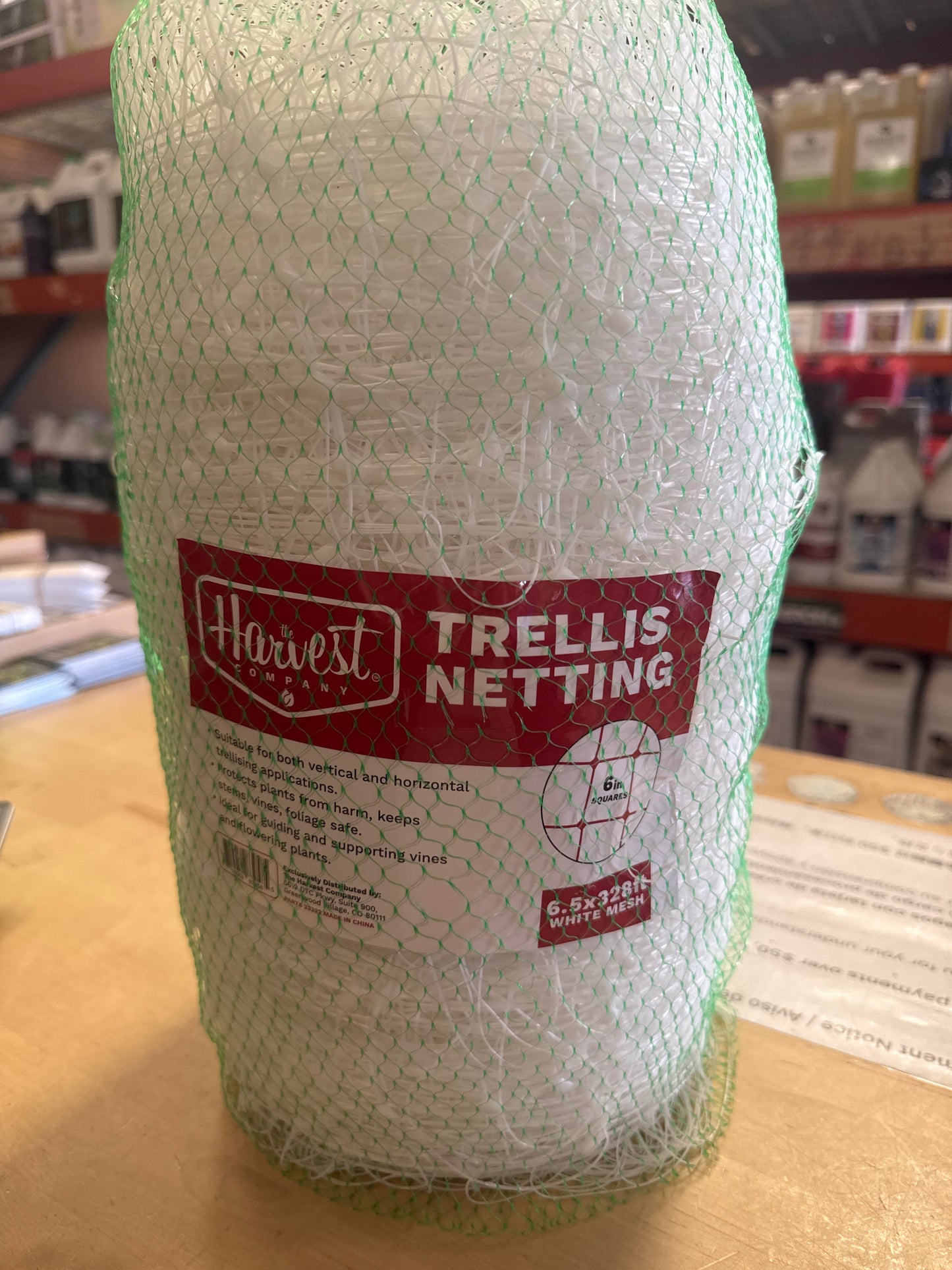 A roll of white trellis netting wrapped in a green mesh protective sleeve, featuring a red and white Harvest Market label, displayed on a wooden counter in a store.